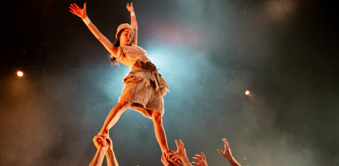 Eine Akrobatin steht mit erhobenen Händen auf den Händen mehrerer männlicher Akrobaten. | © Kalimba